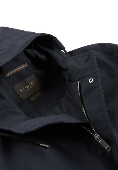 Navy blue jacket with a hood, showing an interior view, features a label, zipper, and a smooth, textured fabric. Minimal accents visible.