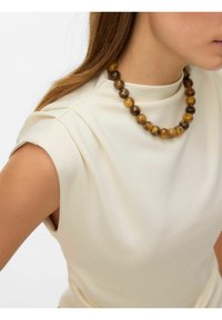 Indumento color crema con collo alto e maniche corte, abbinato a una collana di perline con pietre marroni e gialle.