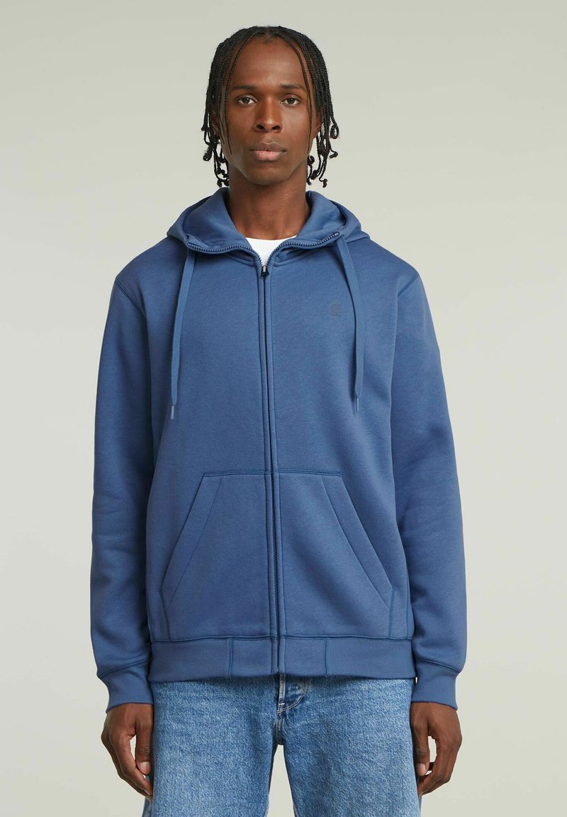Sweatshirt à zip bleu en mélange de coton doux, avec une fermeture éclair avant, une capuche à cordon de serrage et deux poches latérales ; texture unie, coupe décontractée.