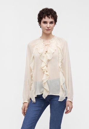 Ung kvinde med kort krøllet hår iført en gennemsigtig cremefarvet bluse med flæser og bindebånd, kombineret med blå jeans, stående mod en hvid baggrund.