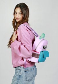 Sac à dos coloré avec un corps rose, une poche inférieure teal et une sangle grise. Présente une fermeture éclair et un logo à l'avant. Design léger.