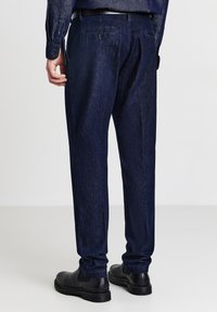 Pantaloni in denim blu scuro con una vestibilità affusolata, dotati di tasche posteriori, sottili pattern di whiskering e orli risvoltati. Completati da calzature nere.