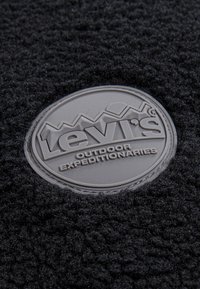Sivi gumeni oznaka s natpisom "Levi's Outdoor Expeditionaries" na crnoj, teksturiranoj tkanini. Oznaka uključuje uzdignuta slova i akcentu planina.