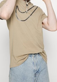 Světle hnědé bavlněné tričko s velkým černým logem Nike v zakřiveném designu. Doplněné o světle modré džíny, které mají volný střih.