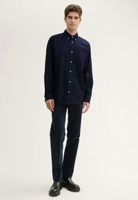 Marineblauw katoenen overhemd met knopen, een borstzak, lange mouwen en witte knopen, gecombineerd met een donkere broek en zwarte schoenen.