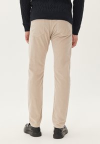 Beige ribfluwelen broek met een recht model, voorzien van twee achterzakken en zwarte instappers, gezien van achteren.