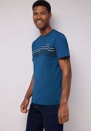 Camiseta de algodón azul con mangas cortas que presenta rayas horizontales en negro y blanco en el pecho. El modelo lleva jeans oscuros.