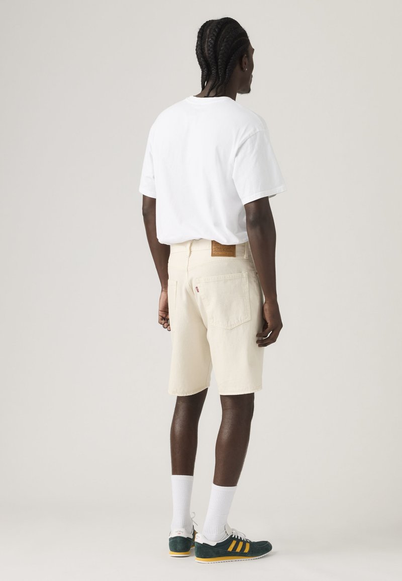 Uomo con capelli intrecciati che indossa una maglietta bianca, pantaloncini beige fino al ginocchio, calzini bianchi e scarpe da ginnastica verdi con strisce gialle, visto di spalle.