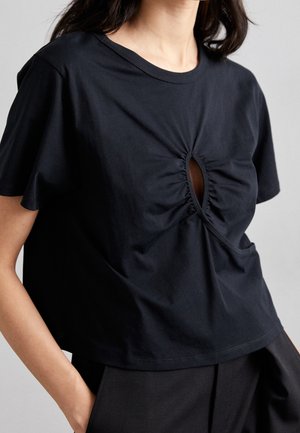 Kobieta ubrana w luźną czarną bluzkę z krótkim rękawem, z marszczonym owalnym wycięciem na wysokości klatki piersiowej, w połączeniu z czarnymi spodniami.