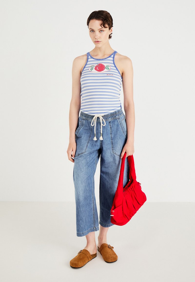 Top sin mangas a rayas en azul y blanco con gráfico de manzana, pantalones anchos de mezclilla azul, zuecos marrones y un bolso acolchado rojo.