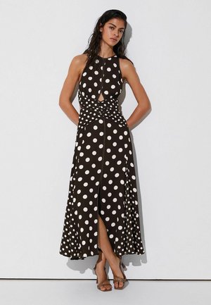 Mujer con el cabello mojado vestida con un vestido midi negro sin mangas con lunares blancos, detalle de corte, abertura frontal y sandalias marrones con tacón.