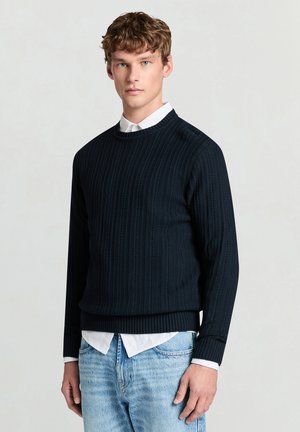 Marineblauer Strickpullover mit Rundhalsausschnitt und geripptem Strukturmuster, mit langen Ärmeln und tailliertem Bund, getragen über einem weißen Hemd mit Kragen.