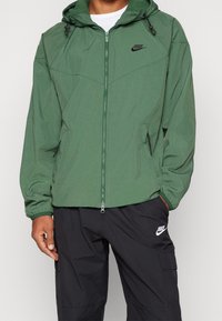 Green lightweight jacket with a hood, zippered front, two side pockets, and black Nike logo. Worn with black pants featuring a small logo.