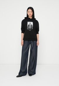 Fekete kapucnis pulóver elülső grafikával, széles szárú sötét farmernadrággal párosítva. A modell egy sima fehér háttér előtt áll.