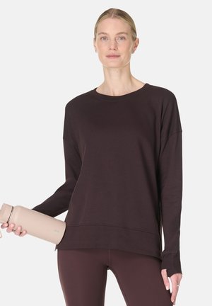 Vrouw met een donker langarmig shirt en legging houdt een beige waterfles vast, staand tegen een eenvoudige lichte achtergrond.