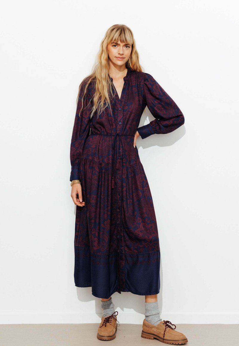 Robe longue à boutons présentant un motif floral rouge foncé sur fond marine. Équipée d'une taille cintrée et de manches longues.