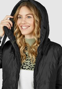 Schwarze Pufferjacke mit Kapuze, ausgestattet mit einem Reißverschluss und gerippten Bündchen. Wird über einem grünen gemusterten Pullover getragen, was einen geschichteten Look unterstreicht.