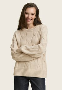 Pull en maille beige avec un motif en torsades, des poignets et un ourlet côtelés. Texture douce, coupe décontractée et encolure ras du cou. Associé à un jean foncé.
