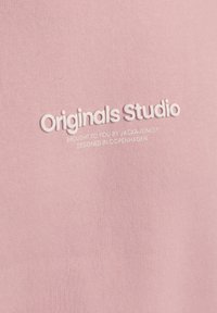 Kerge roosa kangas, millel on sile tekstuur, ja millel on keskel embosseeritud tekst "Originals Studio" ning väiksem tekst, kõik valged.