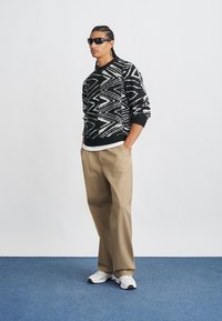 Black and white patterned pullover sweater, beige trousers, and white sneakers. Accessories include black sunglasses and a layered shirt.