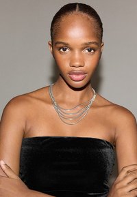 Colier argintiu suprapus purtat pe un gât gol, pe un fundal gri deschis. Modelul poartă un top negru din catifea fără bretele. Textură netedă.