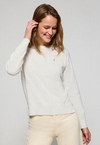 Vit bomullströja med ribbad nederkant och muddar, med en liten broderad logotyp på bröstet. Slät textur, avslappnad passform.