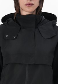 Μαύρο αδιάβροχο μπουφάν με ψηλό κολάρο, κουμπώματα και μπροστινό φερμουάρ. Το ύφασμα φαίνεται λείο και ελαφρώς υφαντό.