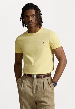 Tricou din bumbac galben, cu guler rotund și mâneci scurte, având un mic logo bleumarin pe piept. Poartă-l cu pantaloni cu un model în carouri și o curea.