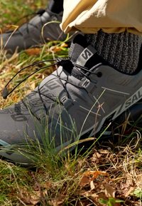 Chaussures d'extérieur grises en matériau texturé, dotées d'un système de laçage sécurisé, d'une semelle en caoutchouc et d'accents de logo ; posées sur de l'herbe et des feuilles.