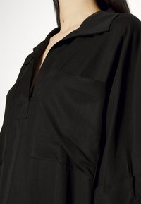 Femme portant une chemise noire à col avec une ouverture en V et deux poches poitrine, visible des épaules jusqu'au milieu du torse.