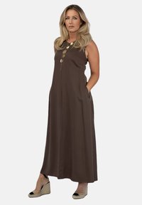 Brązowa, bezrękawnikowa sukienka maxi z guzikami, bocznymi kieszeniami i gładką teksturą. Noszona z czarnymi i beżowymi espadrylami na koturnie.