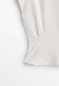 Haut blanc à manches courtes, avec des plis latéraux et une texture lisse. Design simple sans motifs visibles ni accents métalliques.