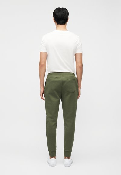 Άνδρας που στέκεται με την πλάτη γυρισμένη, φοράει λευκό t-shirt, χακί παντελόνι φόρμας με τσέπη στο πίσω μέρος και λευκά αθλητικά παπούτσια σε απλό φόντο.