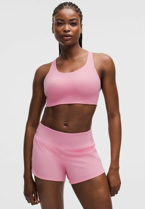 Fit Frau mit geflochtenem Haar, die einen pinkfarbenen Sport-BH und passende Shorts trägt, steht vor einem schlichten hellen Hintergrund.