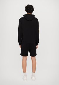 Black hoodie and shorts set, made of soft fabric. Hood attached, with a yellow logo detail on the left sleeve. White sneakers and ankle socks.