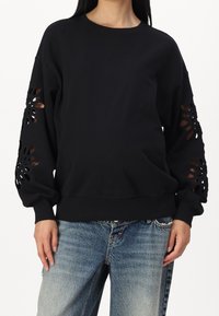 Schwarzer Pullover aus weichem Stoff, mit filigranen ausgeschnittenen Mustern an den Ärmeln, in lockerer Passform und mit gerippten Bündchen. Kombiniert mit blauen Jeans.