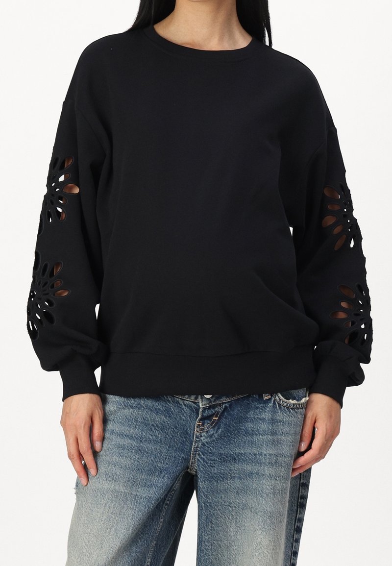 Schwarzer Pullover aus weichem Stoff, mit filigranen ausgeschnittenen Mustern an den Ärmeln, in lockerer Passform und mit gerippten Bündchen. Kombiniert mit blauen Jeans.