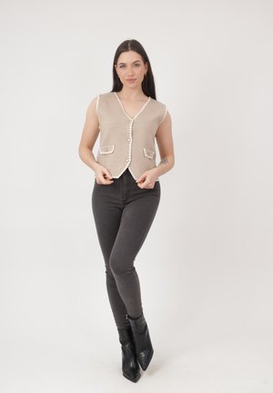 Kobieta stojąca, ubrana w beżową kamizelkę bez rękawów z guziczkami i detalami kieszeni, dopasowane ciemnoszare dżinsy oraz czarne botki na białym tle.