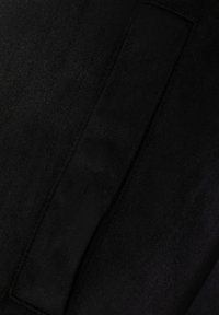 Juodos zomšos audinio arti vaizdas, su lygiu paviršiumi ir stačiakampiu lopu. Dizainas minimalistinis, be matomų raštų ar metalinių detalių.