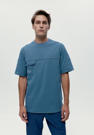 Muž v modrom tričku s krátkym rukávom a náprsným vreckom, tmavomodrých nohaviciach, stojaci pred jednoduchým svetlým pozadím.