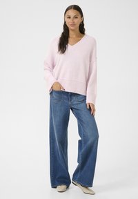 Pullover oversize rosa chiaro con scollo a V e orlo a costine, abbinato a jeans blu a gamba larga e scarpe color crema. Design minimalista.
