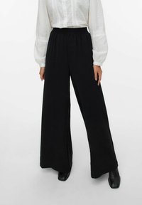 Pantalons larges noirs en tissu doux et extensible avec une taille élastique, associés à un chemisier blanc à manches bouffantes et texturé.