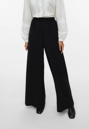 Pantalons larges noirs en tissu doux et extensible avec une taille élastique, associés à un chemisier blanc à manches bouffantes et texturé.