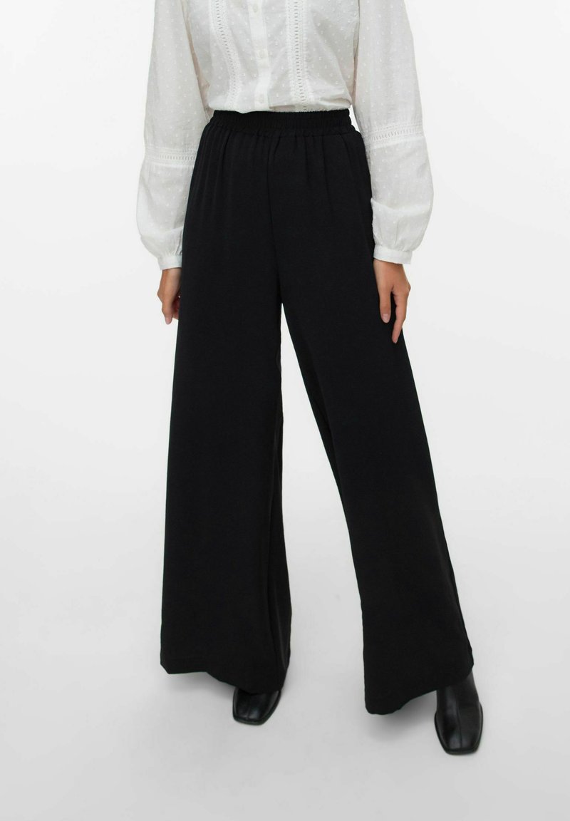 Pantalons larges noirs en tissu doux et extensible avec une taille élastique, associés à un chemisier blanc à manches bouffantes et texturé.