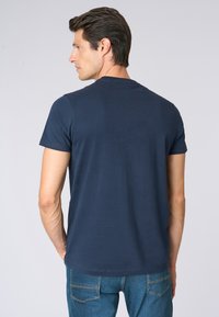 T-shirt in cotone blu navy con collo rotondo e maniche corte. Il retro presenta un dettaglio del logo discreto. Abbinato a jeans in denim blu.