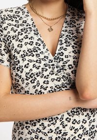 Bluse mit floralem Muster und V-Ausschnitt in Creme, mit schwarzen Blumenmustern. Kurze Ärmel, glattes Material, schichtweise Goldkettchen.