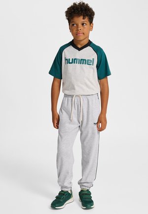 Grijze joggingbroek met zwarte zijstrepen, gecombineerd met een grijs- en tealgekleurde korte mouwen t-shirt met het "hummel"-logo in vetgedrukte letters. Groene sneakers.
