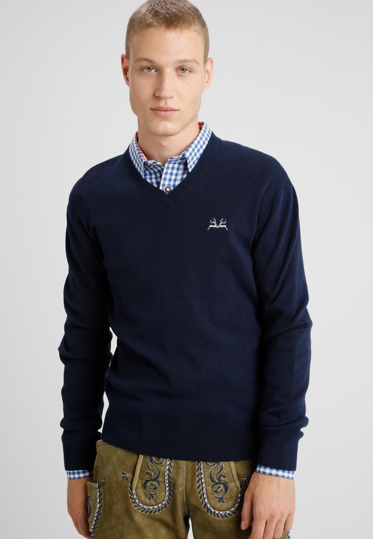 Marineblaues V-Ausschnitt-Pullover aus weichem Material, mit einem kleinen gestickten Design. Über einem karierten blau-weißen Hemd getragen.