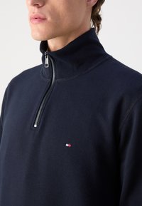 Navy zip-up sweatshirt made of cotton blend with high collar, silver zipper, and small logo on the chest. Smooth texture, casual design.