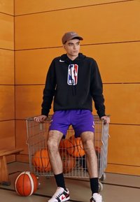 Hanorac negru cu logo-ul NBA, pantaloni scurți mov și adidași albi. Stând lângă un cărucior metalic plin cu mingi de baschet portocalii, pe fundalul unei pereți din lemn.